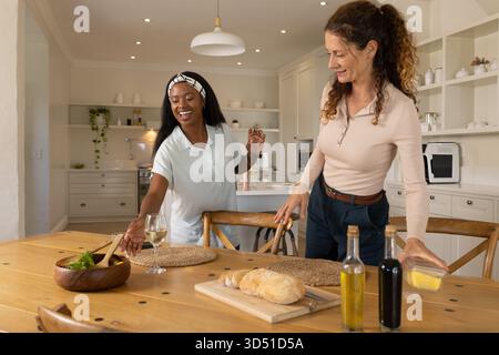 Diverses amies féminines arrangeant la table à manger dans la cuisine moderne, avec saladier et verres à vin Banque D'Images