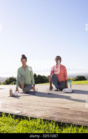 Étirer des amies en tirant les genoux vers des coffres sur une terrasse en bois, avec des tapis de yoga et des bouteilles Banque D'Images