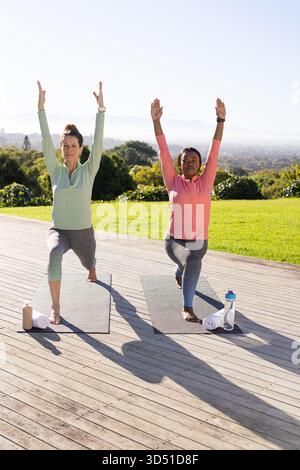 Pratiquer diverses amies féminines tenant des fentes de yoga en avant sur la terrasse en bois, avec des bouteilles d'eau Banque D'Images