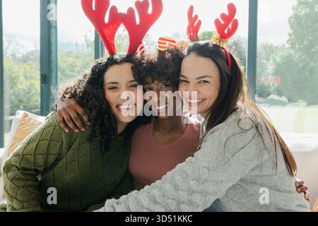 Diverses amies féminines embrassant tout en souriant sur le canapé à la maison portant des bandeaux en bois de renne rouge Banque D'Images