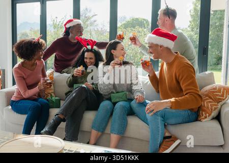 Des amis divers grillent avec des verres à cocktail portant des couvre-chefs festifs autour du canapé dans le salon Banque D'Images