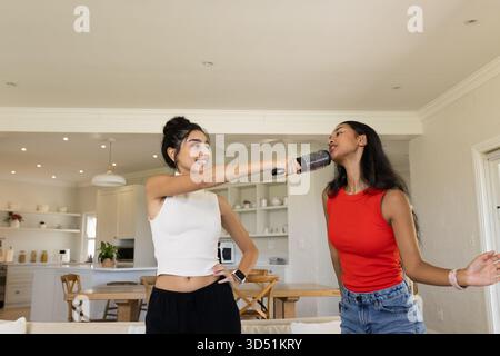 Chanter diverses amies féminines à l'aide du micro brosse à cheveux à l'îlot de cuisine ouvert, avec une table à manger en bois Banque D'Images