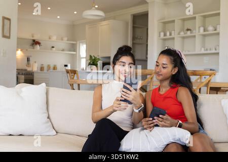 Diverses amies féminines partageant des photos sur smartphones tout en étant assis sur un canapé beige clair dans la cuisine Banque D'Images