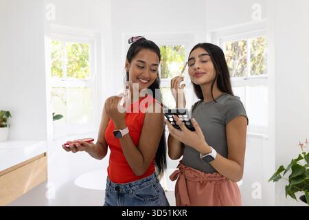 Souriant diverses amies féminines appliquant le maquillage à la vanité de salle de bains moderne, avec des pinceaux et une palette Banque D'Images