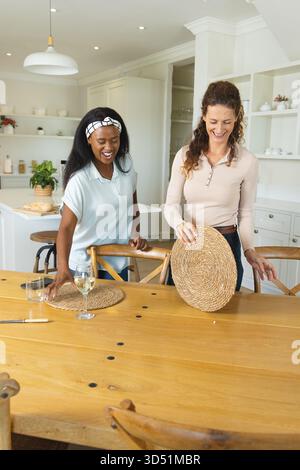 Organiser diverses amies féminines plaçant des sets de table sur la table à manger dans la cuisine avec verre à vin Banque D'Images