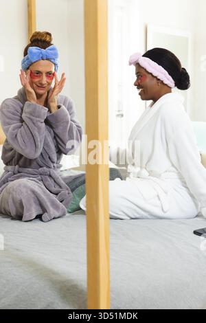 L'application de gel sous les yeux patches diverses amies féminines portant des robes sur le lit dans la chambre, avec miroir Banque D'Images