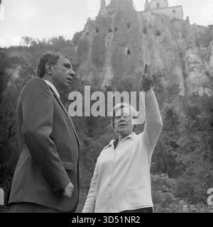 Cette photographie d'archives en noir et blanc de 1975 capture un moment poignant d'une visite guidée à Sviatohirsk (alors Slavyanogorsk), en RSS d'Ukraine. Une femme agit comme guide, pointant avec enthousiasme vers l'église historique de Nicolas, perchée au sommet des falaises de craie massives de la Lavra de Sviatohirsk. À ses côtés, Shutenko, vétéran aveugle, écoute attentivement sa description du célèbre monument. Cette image dépeint magnifiquement un moment personnel de visites et de connexion humaine pour le vétéran de la seconde Guerre mondiale. Banque D'Images