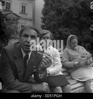 Cette puissante photo d'archives en noir et blanc de 1975 capture un portrait franc et en gros plan du vétéran aveugle Shutenko à Sviatohirsk (alors Slavyanogorsk), en RSS d'Ukraine. Prise lors du 30e anniversaire du jour de la victoire, l'image montre Shutenko parlant avec passion et expression, tenant une cigarette dans un long support. Il est assis sur un banc de parc, tandis que sa compagne et une autre femme (en foulard, lisant un livre) sont assis derrière lui comme un public attentif. En arrière-plan se trouve un grand bâtiment historique. Banque D'Images