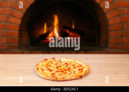 Pizza chaude avec fromage fondu et sauce tomate servie sur une table en bois devant un four rustique en briques avec des flammes de feu ouvert Banque D'Images