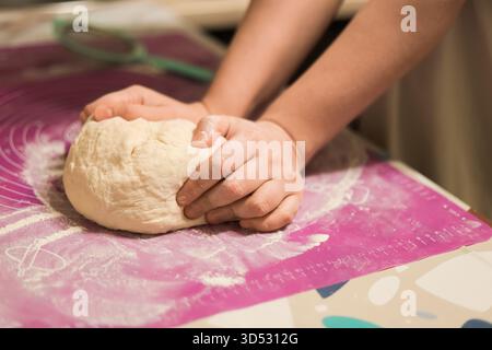Gros plan des mains pressant fermement et pétrissant une boule de pâte fraîche sur un tapis de cuisson en silicone rose saupoudré de farine. Photo de haute qualité Banque D'Images