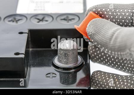 Une main gantée dévisse et retire le capuchon de protection orange vif d'une borne de batterie de voiture, exposant la borne métallique en dessous. Le boîtier de la batterie Banque D'Images
