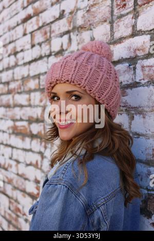 Portrait d'une femme souriante portant une veste en denim et un chapeau en laine debout devant un mur de briques Banque D'Images