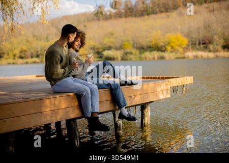 Deux personnes s'assoient ensemble sur un quai en bois, partageant rire et boissons tout en profitant de la vue paisible sur le lac. Banque D'Images