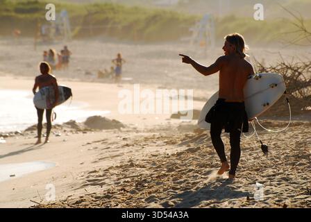 Un jeune homme signale à ses amis tout en portant une planche de surf à la fin d'une journée d'été à la plage de Montauk, New York, sur long Island Banque D'Images