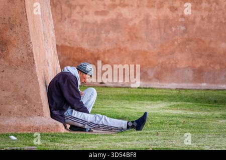 Marrakech, Maroc – 21 avril 2025 : un vieil homme est assis tranquillement contre un vieux mur de terre dans la médina historique. Habillé chaudement dans un bonnet et ja Banque D'Images