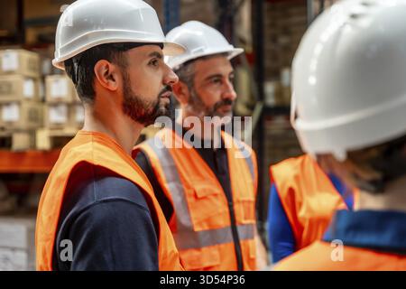 Les employés d'entrepôt portant des gilets réfléchissants et des casques de sécurité écoutent activement pendant une réunion d'équipe, discutent des opérations logistiques et du protocole de sécurité Banque D'Images