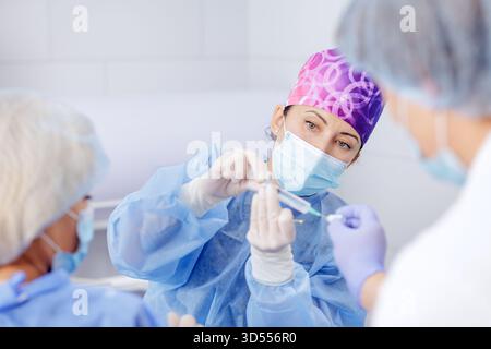 Chirurgien féminin dans le masque protecteur prépare seringue avec médicament pour le patient tandis que les infirmières aident dans la salle d'hôpital stérile brillant Banque D'Images
