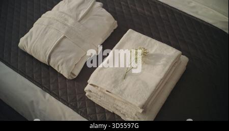 La femme de ménage de l'hôtel en gants blancs plie soigneusement les serviettes blanches fraîches et le peignoir sur le lit soigneusement fait, en plaçant délicat brin de fleur sur le dessus. Concept de propreté, d'entretien et d'expérience client haut de gamme. Banque D'Images