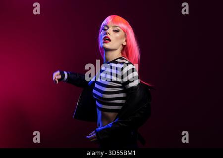 Jeune femme caucasienne avec des cheveux rose vif pose farouchement dans un éclairage au néon dramatique. Cette magnifique mannequin porte un short rayé et une veste en cuir, avec un look rebelle et confiant. Banque D'Images