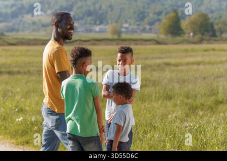 Père sourit en jouant avec ses trois fils dans un champ verdoyant pendant une journée ensoleillée. Banque D'Images