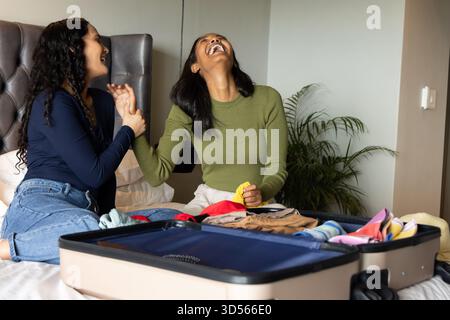 Rire diverses amies féminines emballant des vêtements sur le lit dans la chambre à coucher, avec valise ouverte et plante Banque D'Images