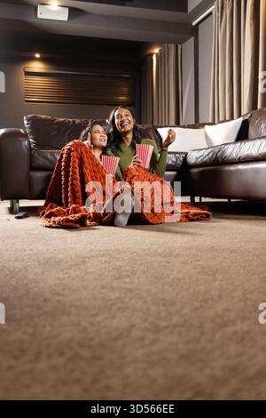 Blottissant diverses amies tenant des baignoires à pop-corn sur le sol du salon, partageant une couverture orange Banque D'Images