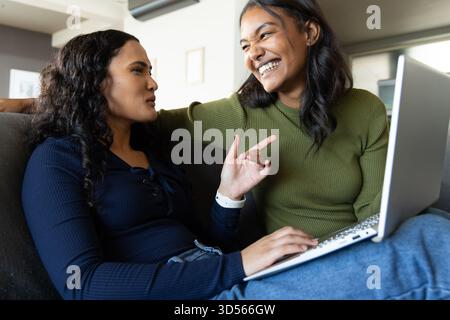Rire diverses amies féminines collaborant sur un canapé sombre à la maison, avec un ordinateur portable argenté Banque D'Images