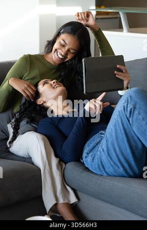 Se prélasser divers couples féminins partageant une tablette noire sur un canapé gris dans un salon moderne lumineux Banque D'Images Se prélasser divers couples féminins partageant une tablette noire sur un canapé gris dans un salon moderne lumineux Banque D'Images