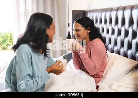 Discutant diverses amies féminines assis sur le lit avec tête de lit touffetée dans la chambre, tenant des tasses blanches Banque D'Images