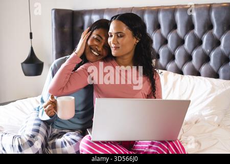 Embrasser diverses partenaires féminines assis sur le lit contre la tête de lit tuftée, avec ordinateur portable et tasse à café Banque D'Images