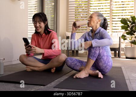 Femme asiatique utilisant un smartphone tandis que la femme plus âgée boit smoothie sur des tapis de yoga à la maison Banque D'Images