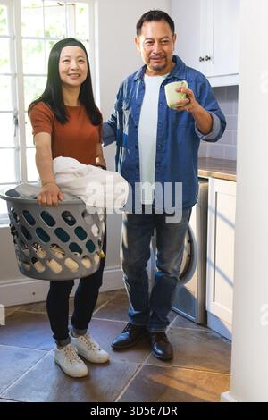 Couple mature faisant la lessive ensemble à la maison, souriant et appréciant le café Banque D'Images