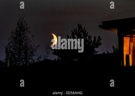Une belle vue d'un morceau de lune dans le ciel sombre, encadrée par des arbres et une maison avec des lumières allumées, ce qui fait sentir calme et invitant. mise au point sélective Banque D'Images