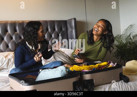 Rire diverses amies féminines emballant la valise ouverte sur le lit à la maison, avec brosse à cheveux et smartphone Banque D'Images