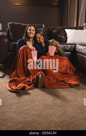Câlin diverses amies féminines sous une couverture orange dans le salon, avec des boîtes de pop-corn Banque D'Images