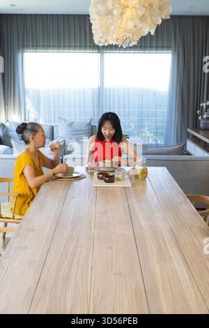 Mère asiatique assise et fille partageant le repas à la table en bois dans le salon, avec des bols en céramique Banque D'Images