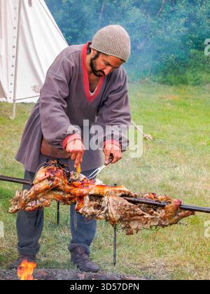 Bøstad, Norvège - 3 août 2023 : un homme en costumes folkloriques médiévaux rôtit un cochon sur une broche pendant le Festival des Vikings au Musée vivant des Vikings Banque D'Images