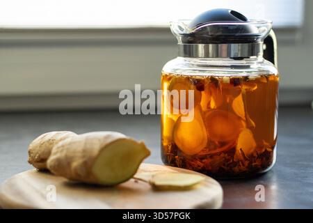 Thé au gingembre chaud dans une théière en verre avec des tranches de gingembre fraîches, sur la table de cuisine grise Banque D'Images