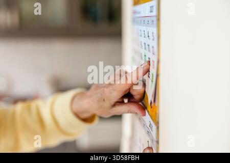 Femme âgée utilisant un calendrier mural dans sa cuisine, concentrez-vous sur le doigt Banque D'Images