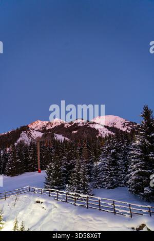 Image verticale en basse lumière de sommets de montagne enneigés au-dessus d'une forêt sombre de conifères, avec une piste de ski et une clôture en bois au premier plan Banque D'Images