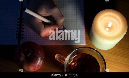 Un étudiant écrit des notes à la lueur des chandelles, dégustant du thé et une pomme pendant le temps d'étude. Banque D'Images