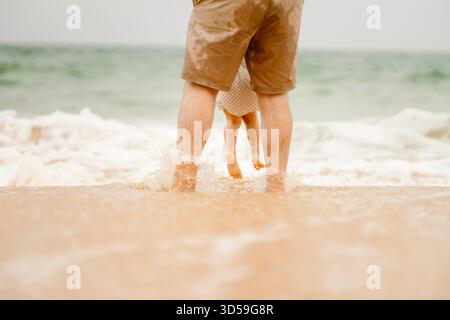 Vue rapprochée des jambes du père et de la fille debout ensemble sur la rive sablonneuse de Praia dos tomates, Algarve, Portugal. De douces vagues de mer touchent th Banque D'Images