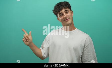 Homme pointant le doigt vers la gauche avec la main droite dans un studio sarcelle portant un t-shirt blanc et souriant, jeune hispanique posture décontractée ; confiance ludique. Banque D'Images