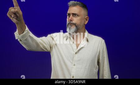 Homme d'âge moyen pointe l'index avec une expression sérieuse dans un studio bleu foncé portant une chemise en lin beige ; autorité. Banque D'Images