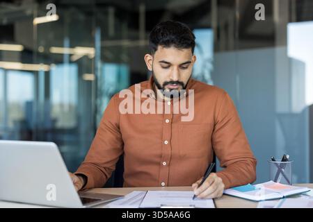 Jeune homme occupé travaillant à son bureau, analysant des documents, remplissant des formulaires et utilisant un ordinateur portable dans un environnement de bureau moderne, représentant la concentration et la productivité de l'entreprise Banque D'Images