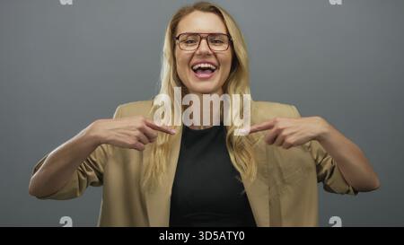 Femme souriant et faisant des gestes à elle-même dans un blazer beige sur un fond gris Uni. Banque D'Images