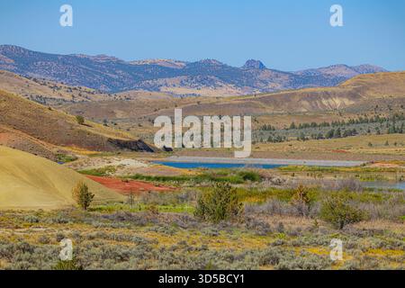 Vaste paysage aride dans Painted Hills Unit avec des collines colorées dorées et rouges, une végétation en arbuste et un petit réservoir bleu Banque D'Images