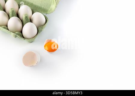 Un carton vert contient plusieurs œufs blancs, tandis qu'un œuf avec une coquille cassée se trouve à côté. La lumière vive améliore les surfaces lisses et les couleurs dans Banque D'Images