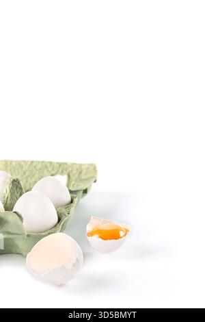 Un carton contient plusieurs oeufs frais, avec un oeuf cassé ouvert à côté, montrant un jaune vif. La scène est placée sur un fond blanc propre Banque D'Images