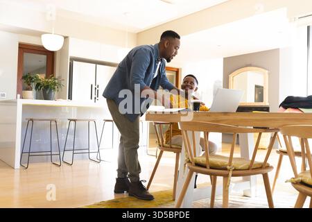 Père afro-américain et fils discutant près de l'ordinateur portable et tasse sur la table à manger dans la cuisine moderne Banque D'Images